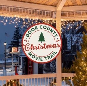 Circular Door County Christmas Movie Trail sign with a green tree, red script text, hanging on a gazebo adorned with string lights at dusk.