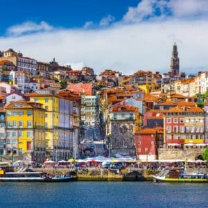 Colorful hillside city by a calm river, with boats at the waterfront and a bright blue sky.