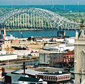 Wide view of a riverfront town with a large steel arch bridge spanning the water and a historic church tower in the foreground.