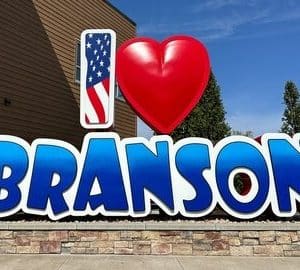 Welcome Branson sign: bold blue BRANSON letters with a red heart and an American flag icon to the left, set on a stone base against a clear sky.