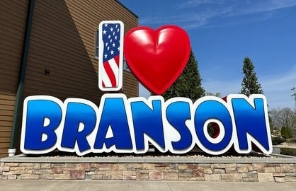 Welcome Branson sign: bold blue BRANSON letters with a red heart and an American flag icon to the left, set on a stone base against a clear sky.