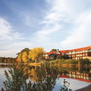Lakeside scene with red-roofed buildings along the shoreline, golden trees, and calm water reflecting the sky in autumn colors.",
