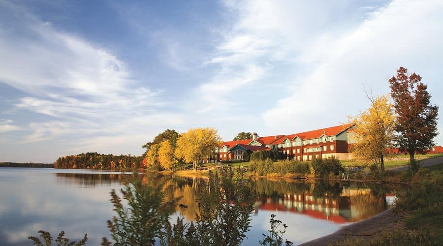 Lakeside scene with red-roofed buildings along the shoreline, golden trees, and calm water reflecting the sky in autumn colors.",