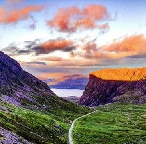 Winding path through a green valley between rocky hills, with a sunset sky of orange clouds above the cliffs on the right.