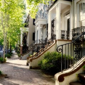 Row of white townhouse facades with ornate wrought-iron stairs and green trees along a sunny sidewalk.